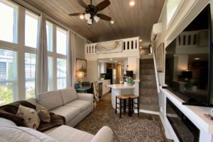 Modern tiny house interior on Just Wandering Lane with large windows, loft bedroom, gray sofa, kitchen, and stairs.