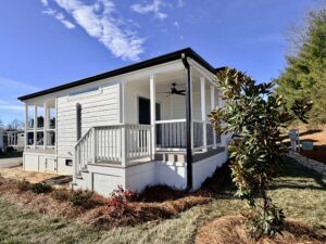 Tiny home with porch, steps, and a fan, surrounded by grass, shrubs, and a blue sky.