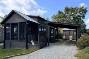 Charming dark gray cabin with porch, carport, and shed on Two Swans Lane, nestled among green trees and gravel driveway.