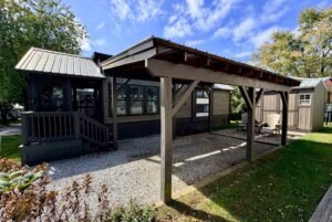A small dark cabin on Two Swans Lane with a covered gravel patio, outdoor chairs, and trees under a blue sky.