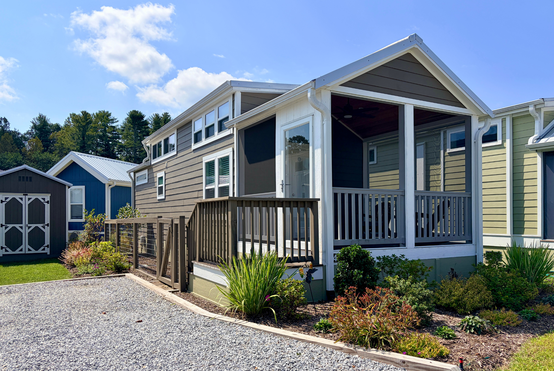 A modern tiny house with a porch on 44 Skipping Stone Lane, surrounded by plants and gravel on a sunny day.