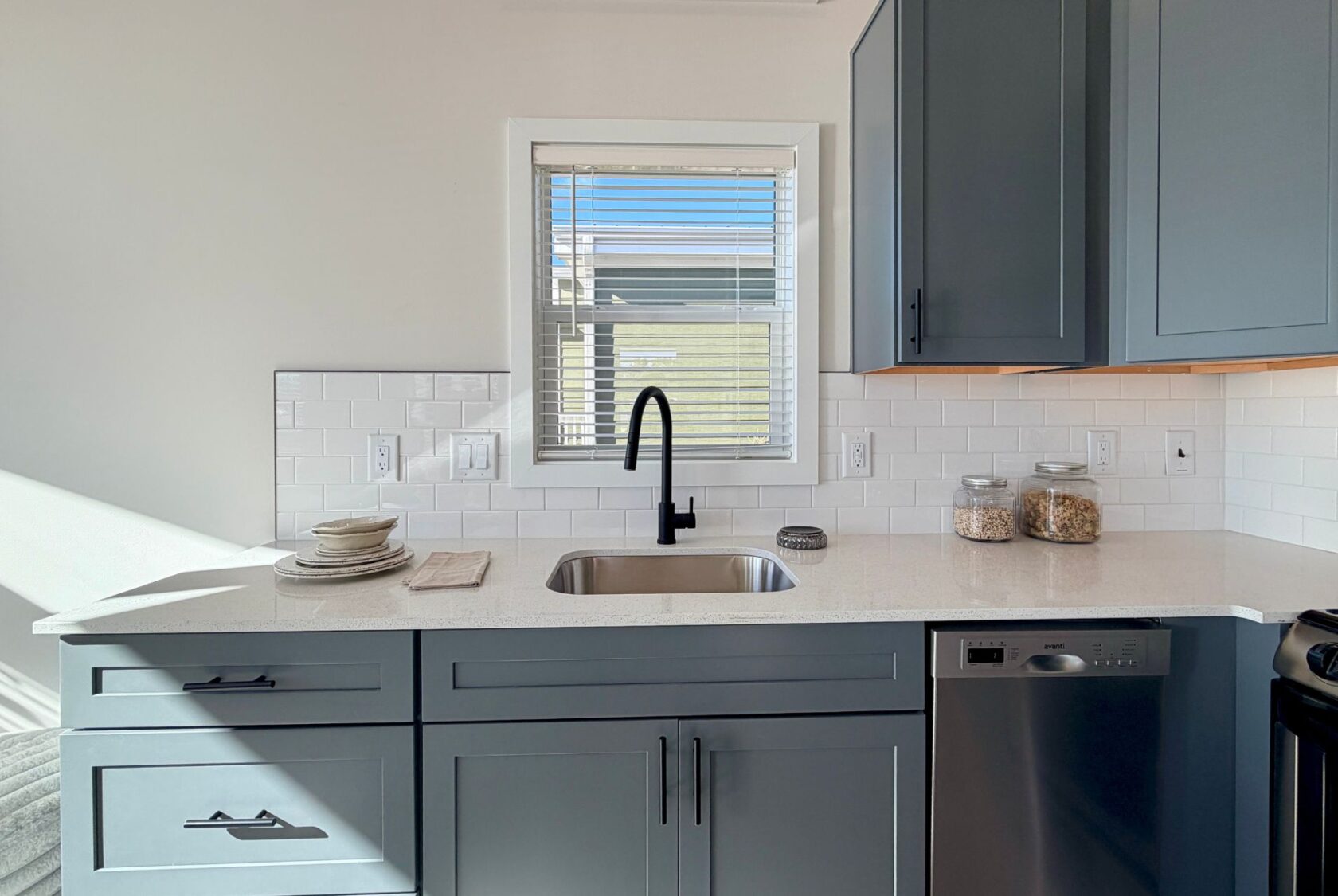 Modern kitchen with blue cabinets, a sink under a window, and jars on a white countertop.