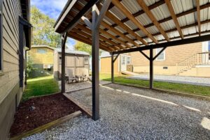 A covered outdoor structure with gravel floor at Two Swans Lane, beside small sheds under a partly cloudy sky.