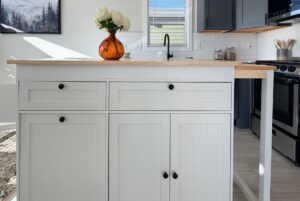 White kitchen island with black knobs, orange vase, modern appliances, and sunlight.
