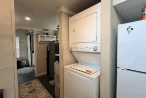 Stacked washer and dryer next to a white fridge in the cozy Two Swans Lane laundry area with shelves and hallway visible.