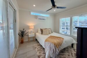 Bright bedroom features a bed, throw blanket, nightstand, lamp, and large sunlit windows.