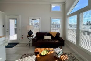 A sunlit living room features a brown sofa, side table, lamp, and coffee table near large windows.