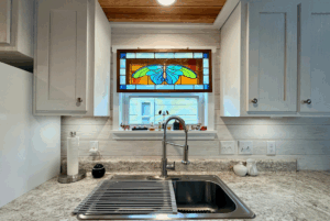 A kitchen sink under a stained glass window on Skipping Stone Lane, with light gray cabinets and marble countertops.