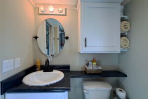 Charming small bathroom at Two Swans Lane with a round mirror, cabinet, shelves, and a basket above the toilet.