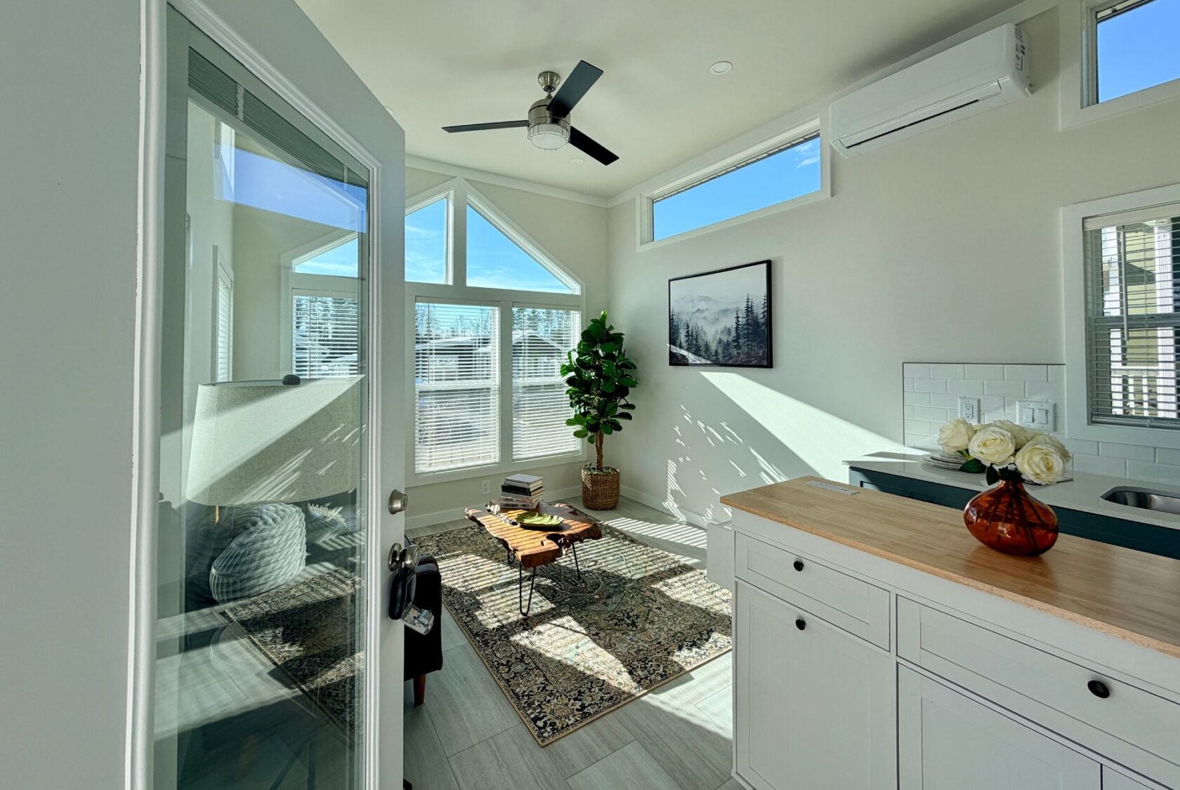 Bright, modern tiny home interior with large windows, a plant, rug, and kitchen counter flowers.