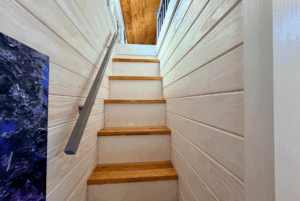 Narrow wooden staircase with white paneled walls leads to a bright space at 44 Skipping Stone Lane with a wooden ceiling.