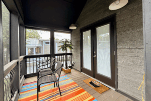 Screened porch at Two Swans Lane with chairs, a striped rug, and doormat, overlooking houses and trees.