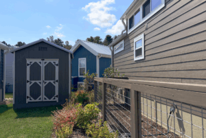 Three small modern homes with gardens line Skipping Stone Lane, enclosed by a wire fence under a partly cloudy sky.