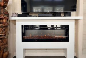 A white electric fireplace from Two Swans Lane glows beneath a TV, beside a wooden statue and a small owl figurine.
