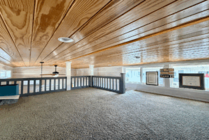 Loft room at Skipping Stone Lane with wood-paneled ceiling, carpeted floor, windows, and a railing overlooking below.