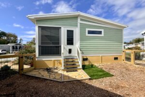 Small green and beige house with steps on Strolling Easy Lane, fenced yard, and blue sky with scattered clouds above.