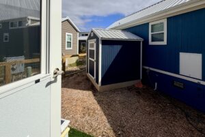 View of a small blue shed beside a blue house on Strolling Easy Lane, seen from an open door on a sunny day.