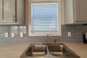 Kitchen sink under a window with closed blinds, gray cabinets, and a Strolling Easy Lane subway tile backsplash.