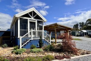 A small modern blue tiny house on Easy Lane with a porch, garden, and wooden carport under a partly cloudy sky.