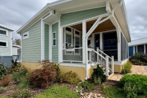 Green and white house with a small front porch, landscaped garden, and Strolling Easy Lane under a cloudy sky.