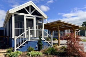 A small blue cottage on Easy Lane with a screened porch, front steps, and a carport beside a landscaped garden.