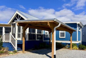 Blue house with white trim on Easy Lane, wooden carport, gravel driveway, and a partly cloudy sky.