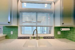 Kitchen sink with green tile backsplash, window above with blinds, and a Strolling Easy Lane vibe throughout.