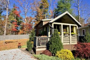 Tiny house on Highland Hollow Lane with a front porch, nestled among autumn trees and lush garden plants.