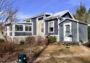 A small, modern blue-gray house with white trim, surrounded by a yard and bare shrubs under a blue sky.