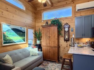 Cozy wooden living room with plants, a sofa, wall clock, painting, and bright natural light from windows.