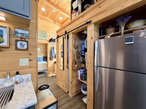 Cozy wooden kitchen with a stainless steel fridge, pantry, and view into a bedroom with a blue bedspread.
