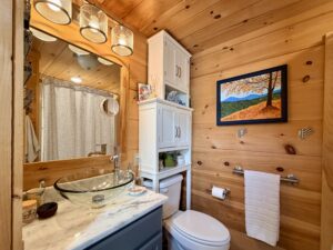 Cozy wooden bathroom with vessel sink, storage above toilet, and framed landscape painting on the wall.