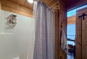 Shower with patterned curtain in a wood-paneled bathroom, towel hanging on wall, doorway to another room.