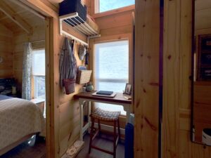 Small wooden desk with a stool by a window, surrounded by shelves and cozy decor in a sunlit cabin room.