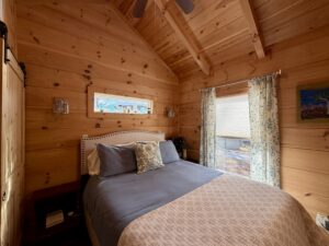 Cozy wooden bedroom with a neatly made bed, floral curtains, and warm natural light from two windows.