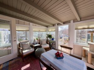 A sunroom with cozy seating, a table with apples, houseplants, and sunlight streaming through many windows.