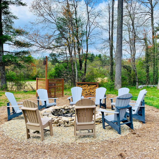 A circle of Adirondack chairs surrounds a fire pit, offering simple life comforts in a sunny wooded backyard.
