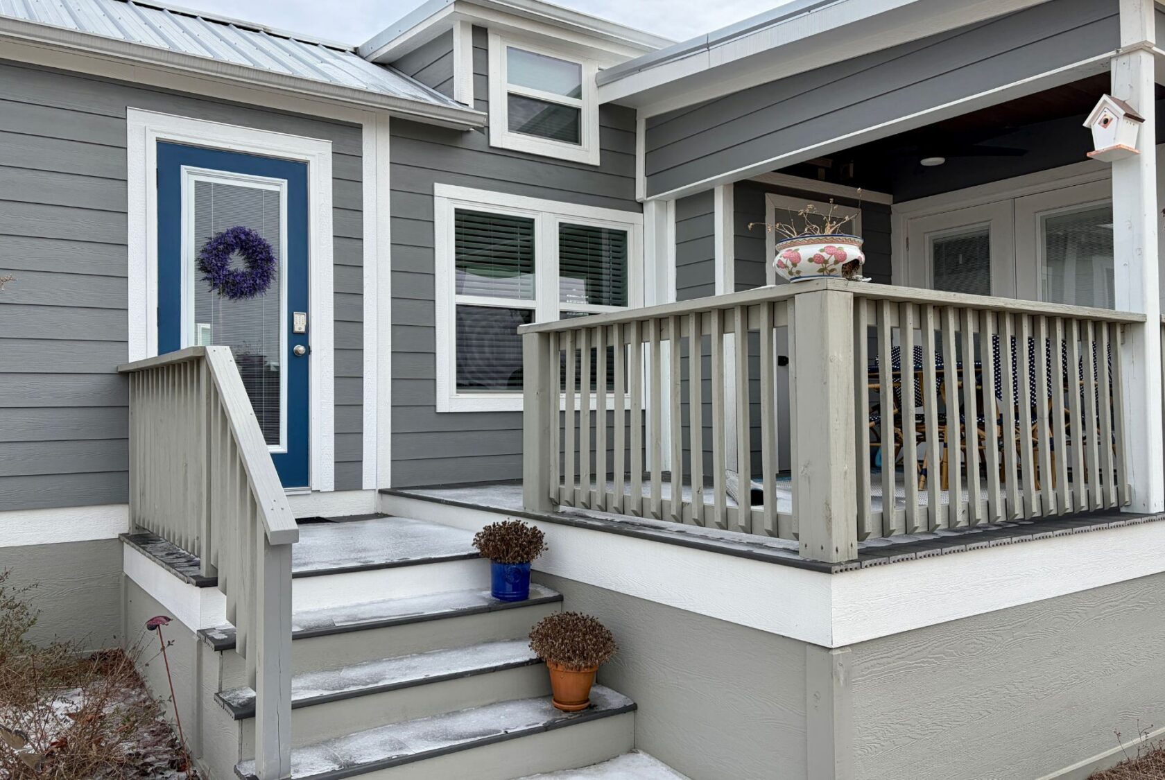 Gray house with blue door on Mount Meadow Lane, small porch, dried plants in pots, light snow on steps and railing.