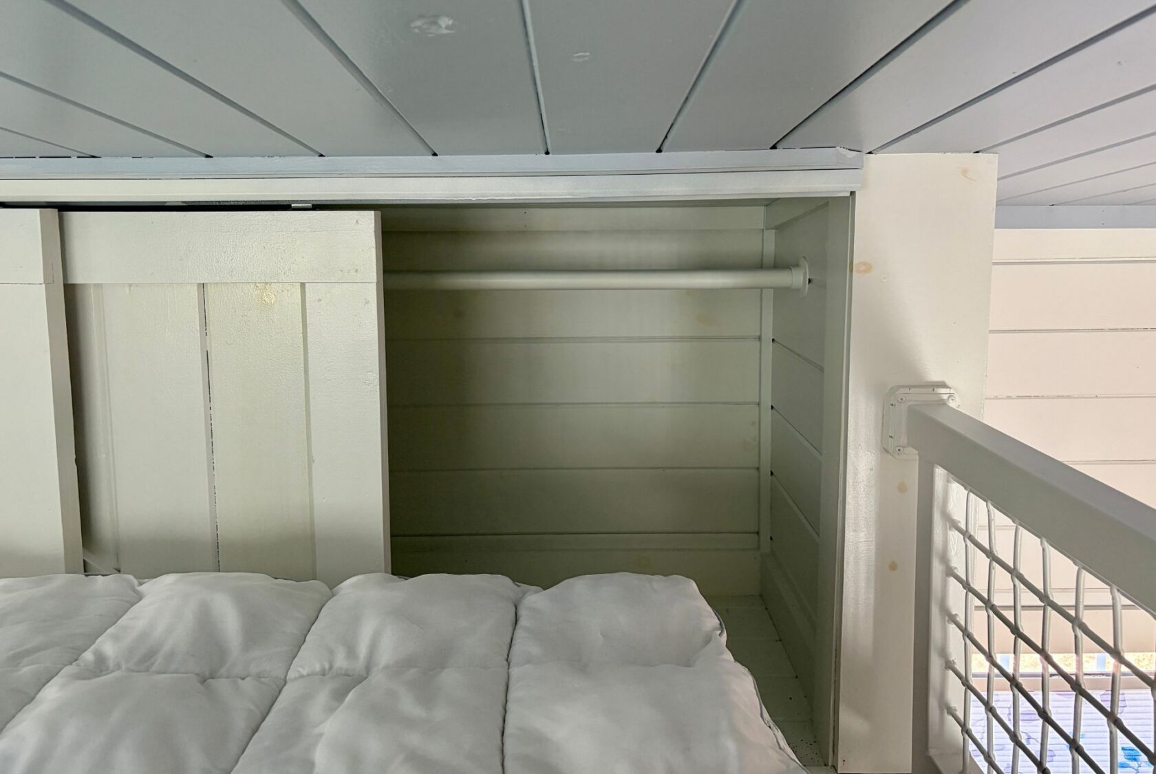 A small, empty white Empire Lane closet with a hanging rod sits beside a bed with a white comforter under a low ceiling.