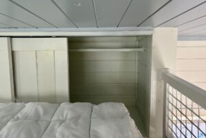A small, empty white Empire Lane closet with a hanging rod sits beside a bed with a white comforter under a low ceiling.