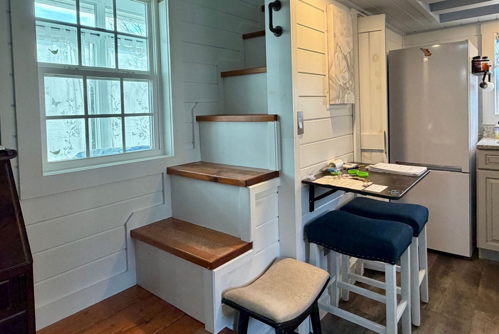 Wooden stairs beside a window, with a small Empire Lane fold-out table and stools near a refrigerator in a cozy kitchen.