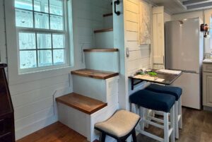 Wooden stairs beside a window, with a small Empire Lane fold-out table and stools near a refrigerator in a cozy kitchen.