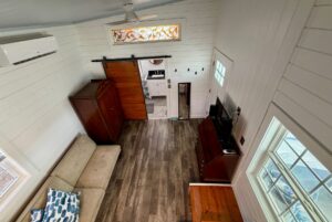 A cozy, white-paneled Empire Lane living room with a sofa, armoire, TV, and a view into a small bathroom.