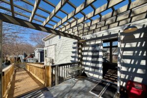 Wooden deck with pergola casting shadows, table, and ramp leading to 139 Drifting Brook under a clear blue sky.