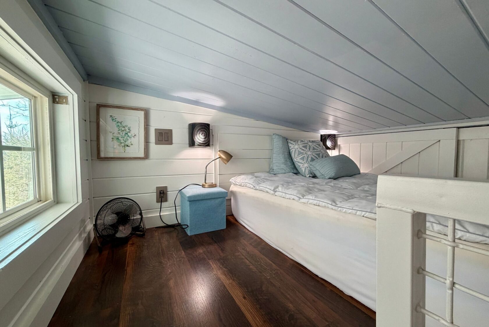 Cozy Empire Lane loft bedroom with a bed, blue decor, small lamp, fan, and window letting in natural light.