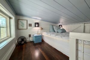 Cozy Empire Lane loft bedroom with a bed, blue decor, small lamp, fan, and window letting in natural light.