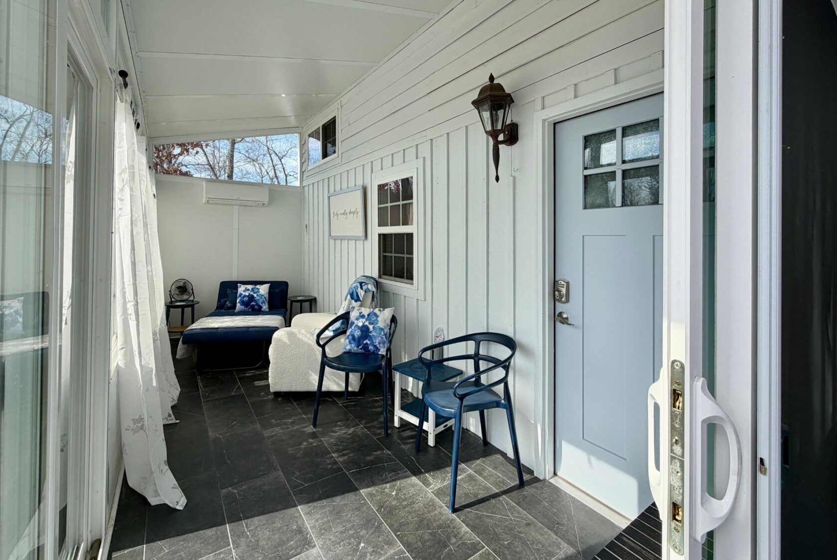 Enclosed porch on Empire Lane with blue door, two blue chairs, sofa, white walls, and sun-filled large windows.