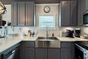 Modern Mount Meadow Lane kitchen with gray cabinets, farmhouse sink, coffee makers, and a clock above window blinds.