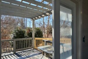 View from a glass door to a wooden deck on Drifting Brook Lane, with railing, pergola, and views of trees and bushes.