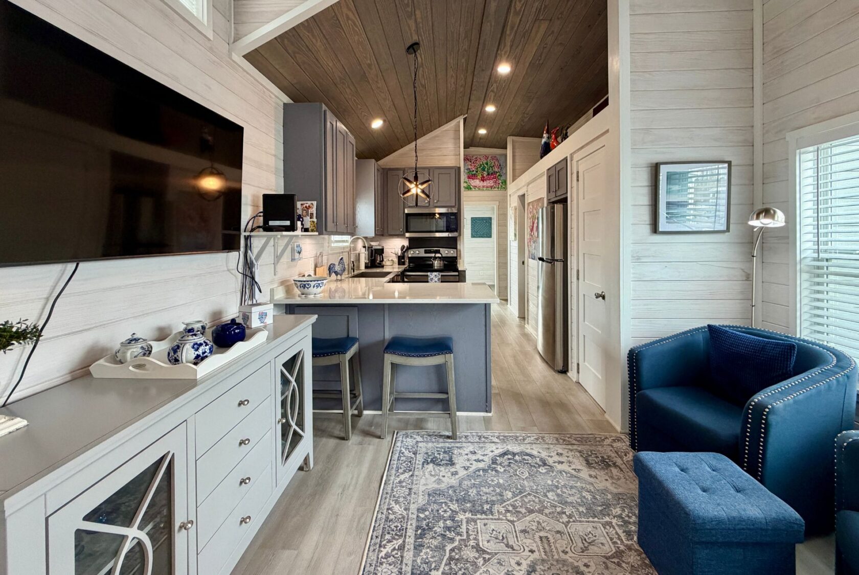 Modern Mount Meadow Lane tiny home interior with kitchen, island seating, blue armchair, and wall-mounted TV.